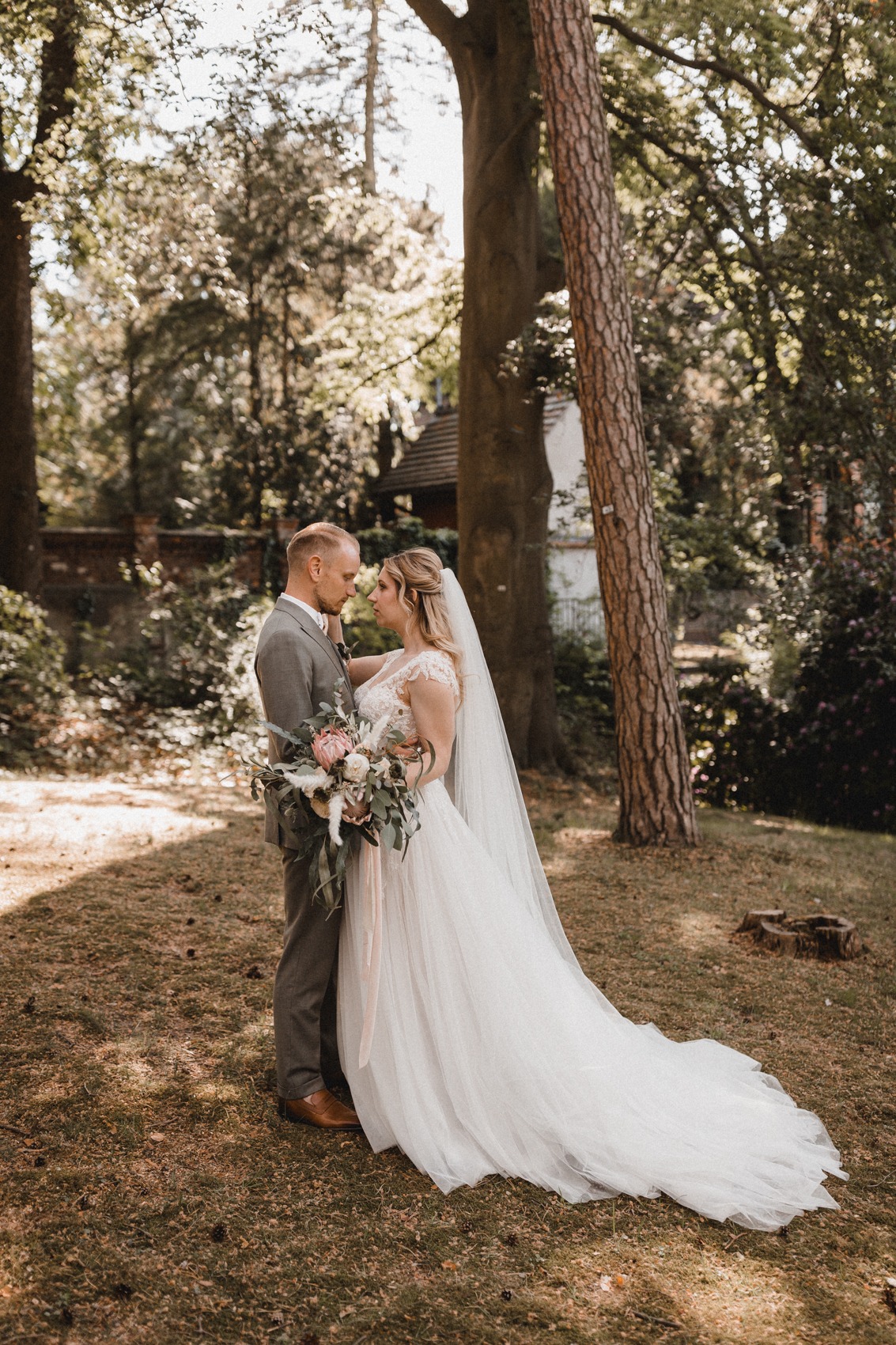 Das verliebte Brautpaar. Der Bräutigam trägt einen graugrünen Zweireiher und die Braut ein Brautkleid in A-Linie mit ausgestelltem Tüllrock und langer Sleppe