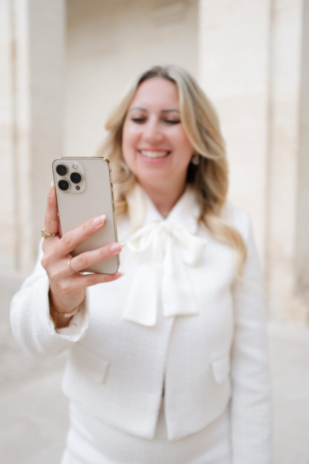 Frau mit Smartphone, lächelnd, in eleganter weißer Jacke, im urbanen Umfeld.