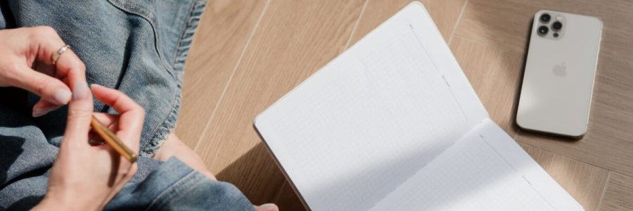 Person sitzt auf Holzboden mit Notizbuch, Smartphone und Kaffee, kreative Arbeitsumgebung.