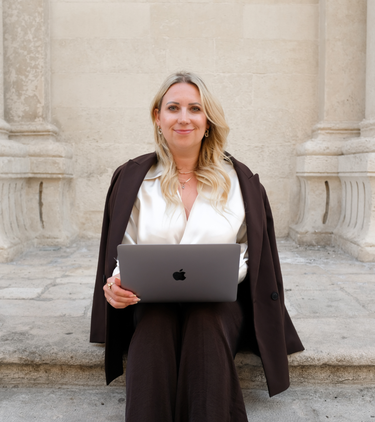 Eine Frau mit Laptop sitzt vor historischen Säulen, professionell und selbstbewusst.